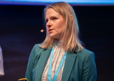 Eine Frau mit blonden Haaren, die einen blaugrünen Blazer und ein Headset-Mikrofon trägt, steht auf der Bühne und schaut aufmerksam zu. Um ihren Hals hängt ein blaues Schlüsselband, und sie hält einen kleinen Gegenstand in der Hand.