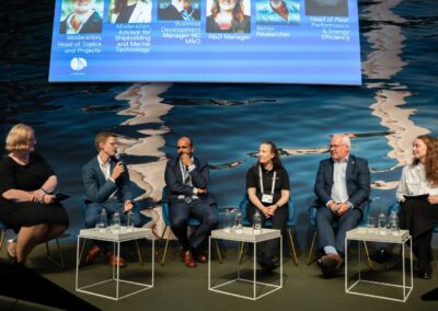 Sechs Diskussionsteilnehmer sitzen mit Mikrofonen auf der Bühne und diskutieren vor einer Leinwand, auf der ihre Rollen und Namen angezeigt werden; hinter ihnen werden Wasserbilder projiziert. Vor ihnen stehen fünf kleine Tische mit Wasserflaschen.