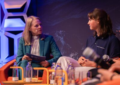 Zwei Frauen sitzen auf der Bühne und unterhalten sich. Die eine hält Notizen in der Hand und trägt einen blaugrünen Blazer, die andere ein marineblaues Poloshirt. Auf einem Tisch vor ihnen stehen Mikrofone und Wasserflaschen. Der Hintergrund ist dunkel und mit abstrakten Mustern verziert.