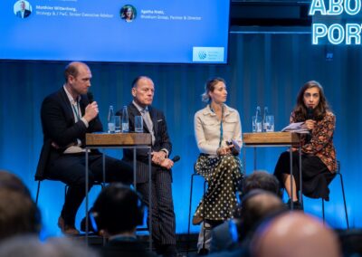 Vier Diskussionsteilnehmer sitzen mit Mikrofonen und Wasserflaschen auf der Bühne und diskutieren auf einer Konferenz zum Thema Häfen, während das Publikum zusieht und auf einem blau beleuchteten Hintergrund ihre Namen und Titel zu sehen sind.
