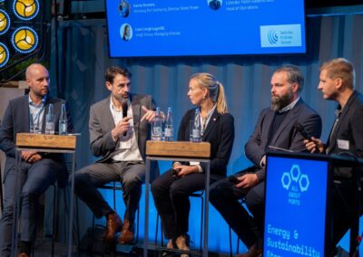 Fünf Personen in Geschäftskleidung sitzen auf einer Podiumsbühne und diskutieren. Ein Mann spricht in ein Mikrofon, während die anderen zuhören. Auf einem blauen Bildschirm neben ihnen sind Sechsecke und die Worte „Energie & Nachhaltigkeit“ zu sehen.