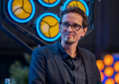 Ein Mann mit Brille und dunklem Blazer sitzt unter blauen und gelben Lichtern und blickt mit nachdenklichem Gesichtsausdruck zur Seite. Der Hintergrund ist sanft unscharf und weist kreisförmige, beleuchtete Formen auf.