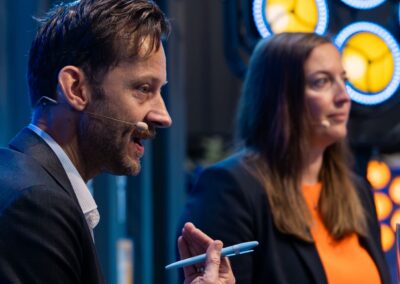 Ein Mann und eine Frau in Business-Kleidung sprechen bei einer Veranstaltung in ihre Headsets. Der Mann hält einen Stift in der Hand und gestikuliert beim Sprechen. Im Hintergrund sind helle, runde Lichter zu sehen.
